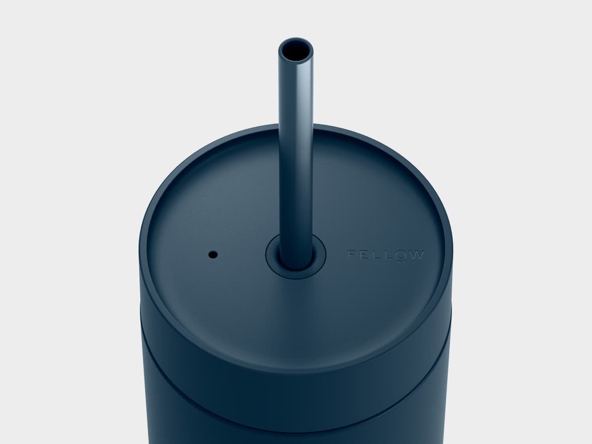 A top-down, close-up view of a dark blue travel mug with a textured finish. A black straw extends from the center of the lid, which also features a small circular vent hole to the left and an embossed "FELLOW" logo to the right. The background is a plain light grey.