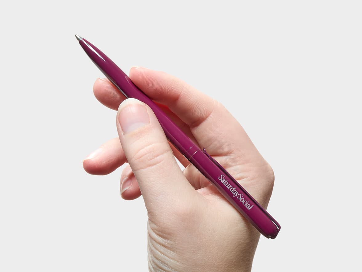 A hand holds a mulberry pen with "SaturdaySocial" written on its side.