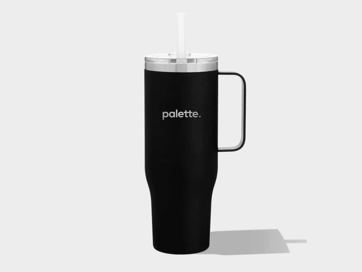 A tall, matte black insulated tumbler with a clear lid, a white straw, and an integrated handle on the right side. The word "palette." is printed in white on the front. It stands upright on a plain light gray surface, casting a shadow to its right.