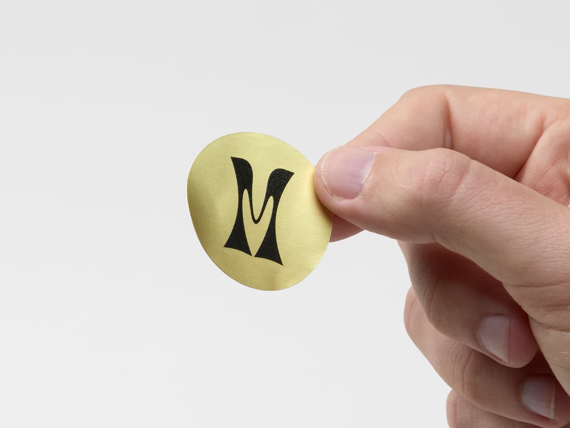 A hand holding a round gold metallic sticker with black customization.