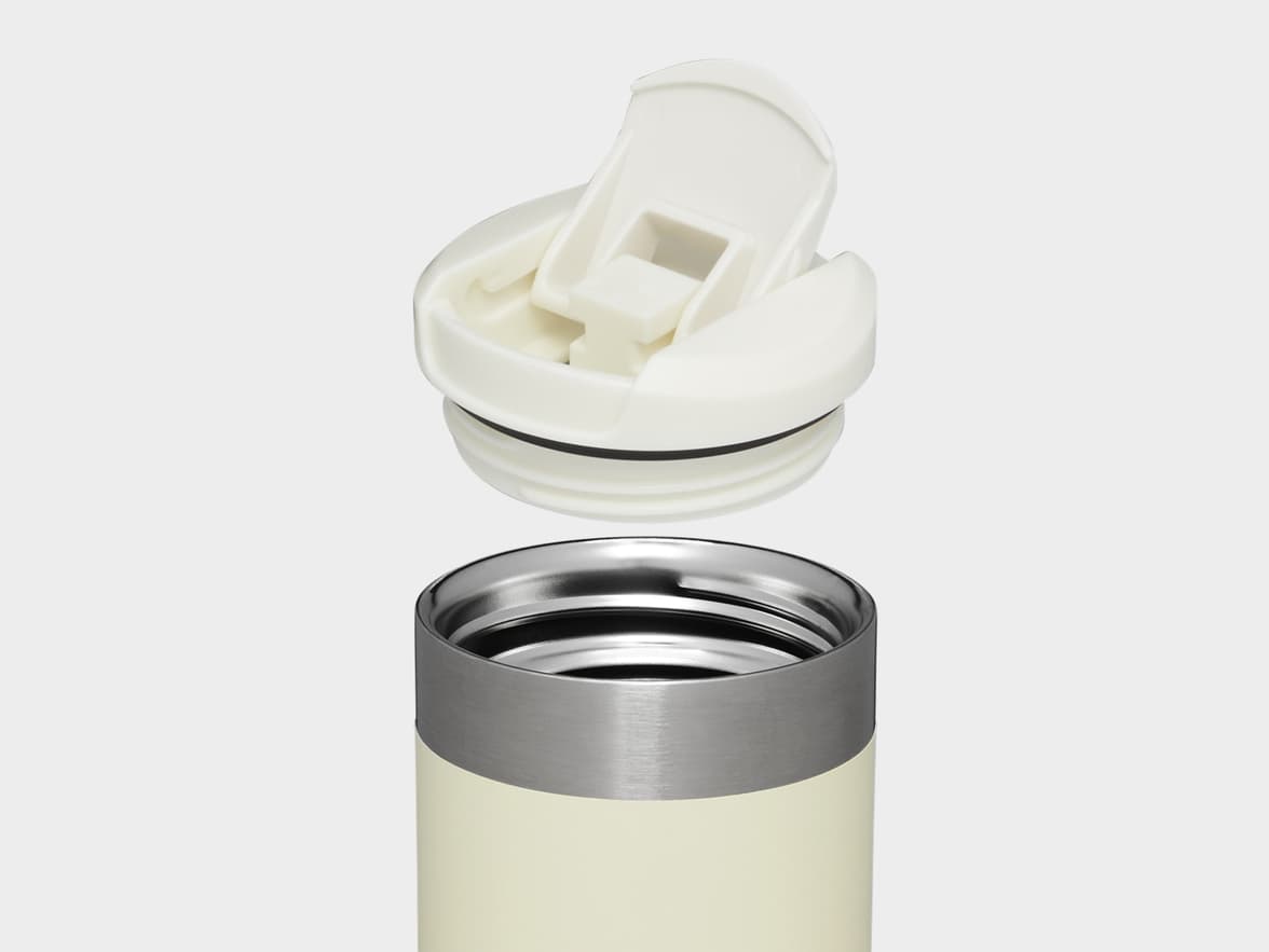 A cream-colored travel mug with a stainless steel rim and its white flip-top lid detached above it.