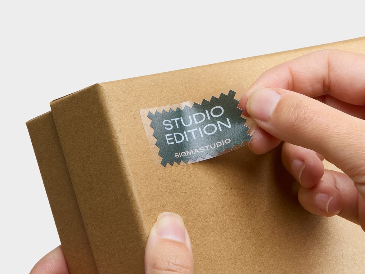 A hand applies a clear sticker with "STUDIO EDITION SIGMASTUDIO" to a brown box.