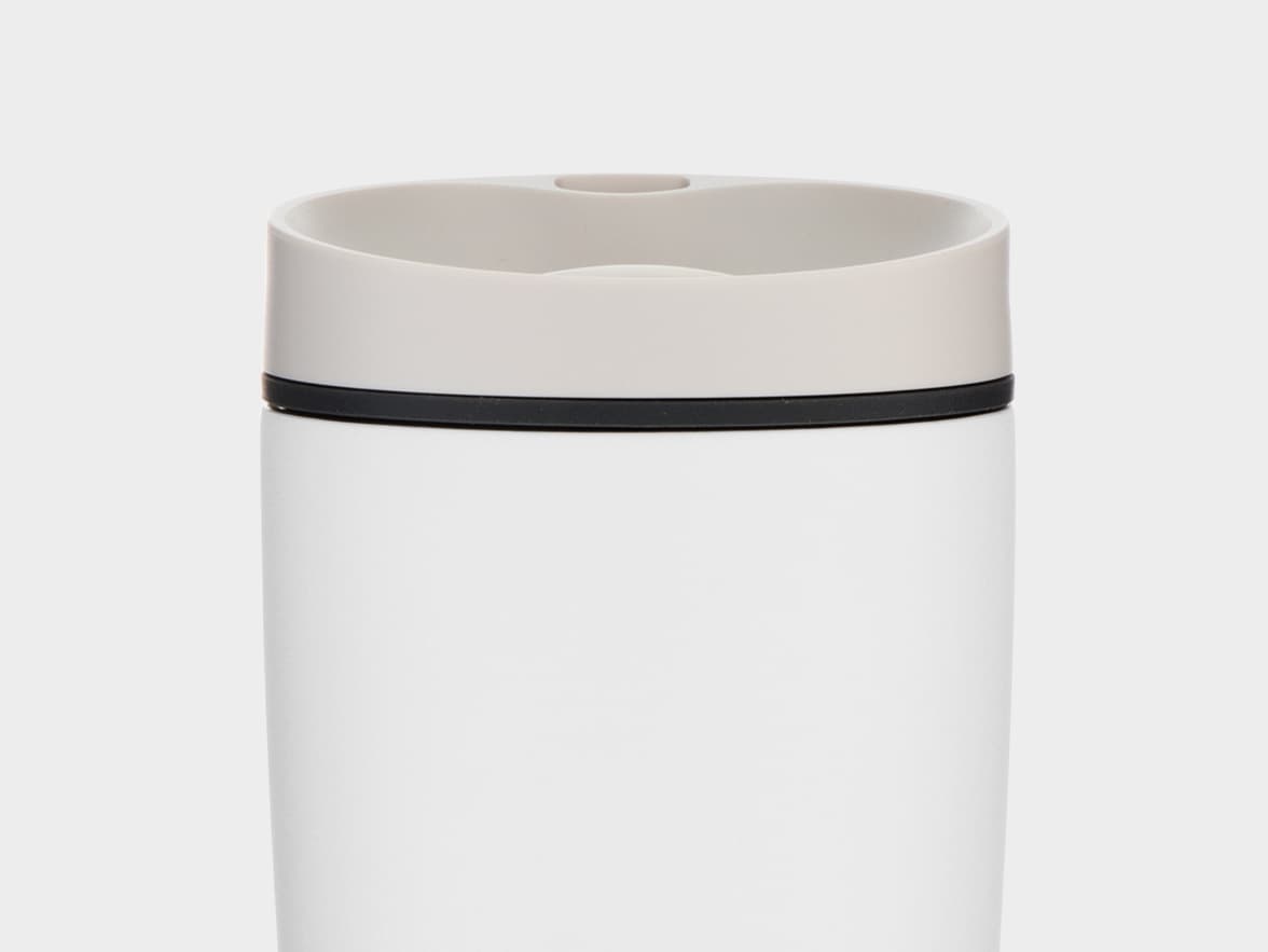 A close-up, eye-level shot of the top portion of a white, cylindrical travel mug with a light grey lid. A thin black band separates the lid from the white body of the mug. The background is a plain, light grey.
