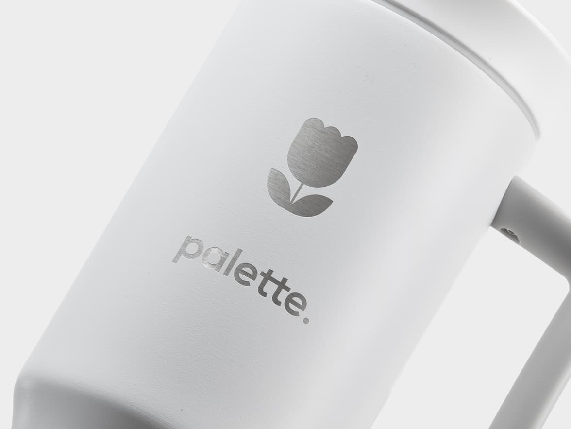 A close-up, angled shot of a white travel tumbler with a matte finish. A silver, metallic logo of a stylized tulip is visible, with the word "palette." below it. The background is light grey.