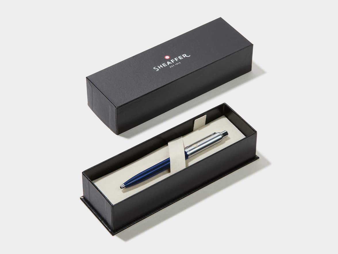 A blue and silver pen rests in an open black Sheaffer gift box with the lid displaying 'SHEAFFER EST. 1913'.