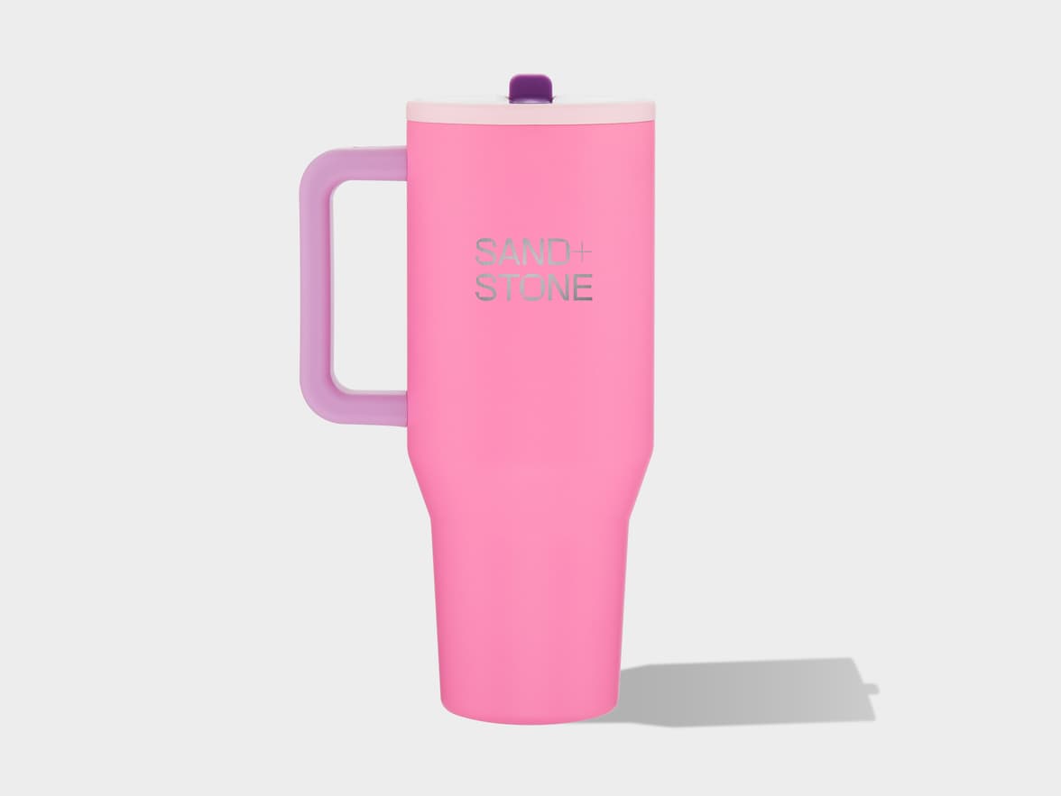 A pink insulated tumbler with a purple handle and a light pink lid topped with a purple cap. The words "SAND + STONE" are printed vertically in silver lettering on the side. The tumbler casts a shadow on a plain light grey background.