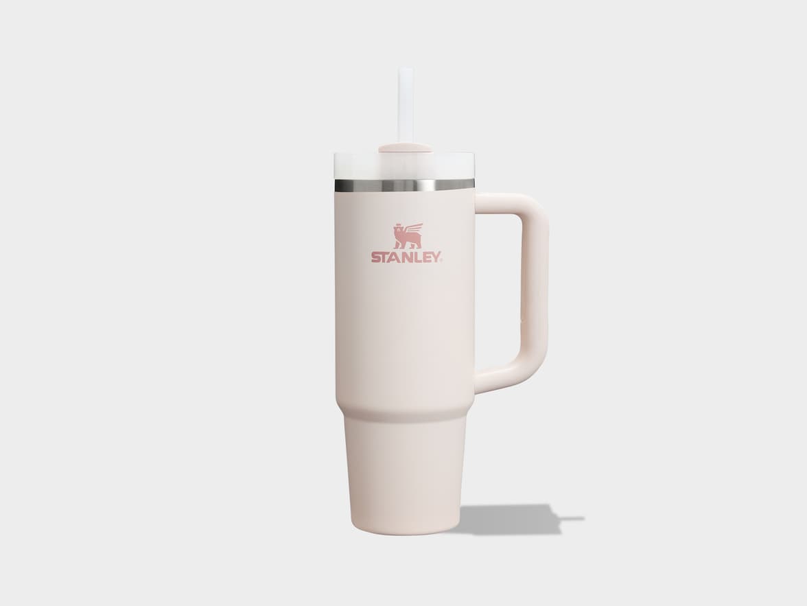 A light pink Stanley Quencher FlowState H2.0 insulated travel tumbler with a clear straw is centered on a light gray surface, casting a subtle shadow to the right. The Stanley logo is visible on the front of the cup, and it features a side handle.