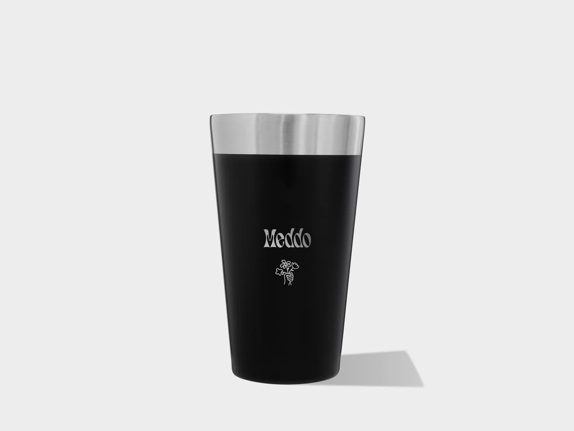 A black and silver tumbler with "Meddo" and a floral design printed on it, standing on a light grey surface with a shadow.