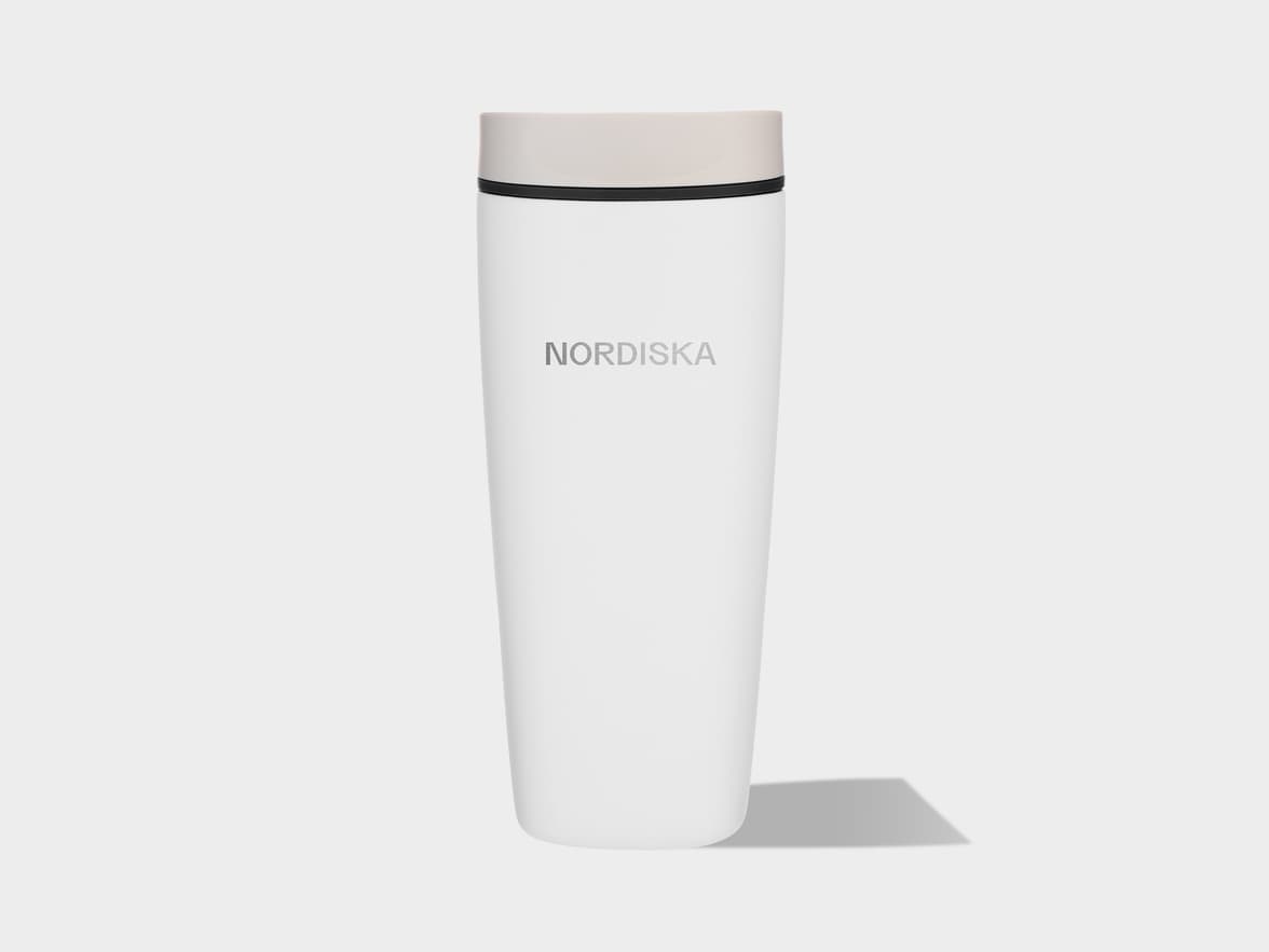 A white cylindrical travel mug with a light beige screw-top lid and a thin black band below the lid. The word "NORDISKA" is printed vertically in light gray letters on the front of the mug. The mug stands upright on a light gray surface, casting a subtle shadow to its right.