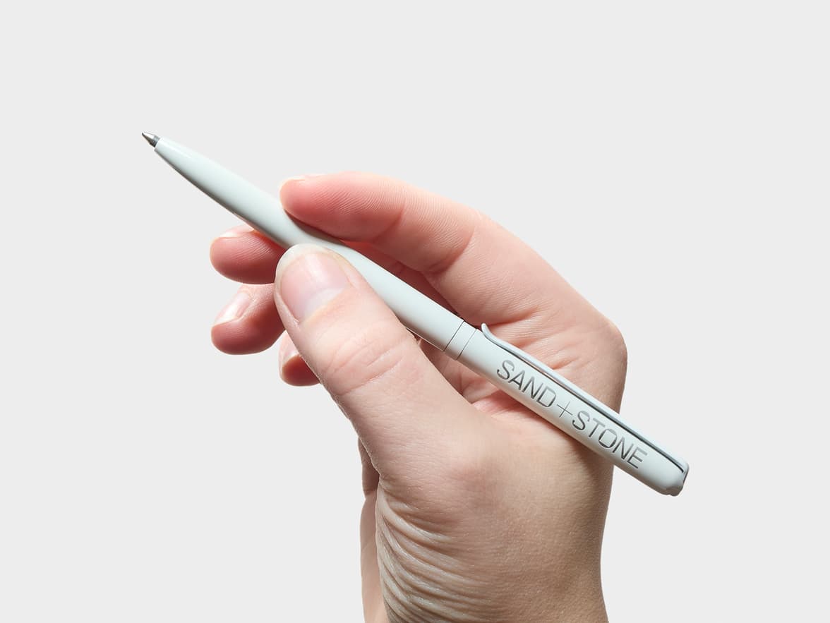 A hand holds a light-colored pen with "SAND + STONE" branded on its clip.