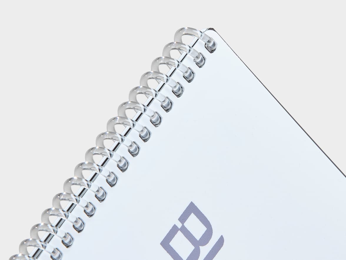 Close-up of a white spiral-bound notebook with a clear plastic coil, showing a partial light purple "B" logo on the cover.