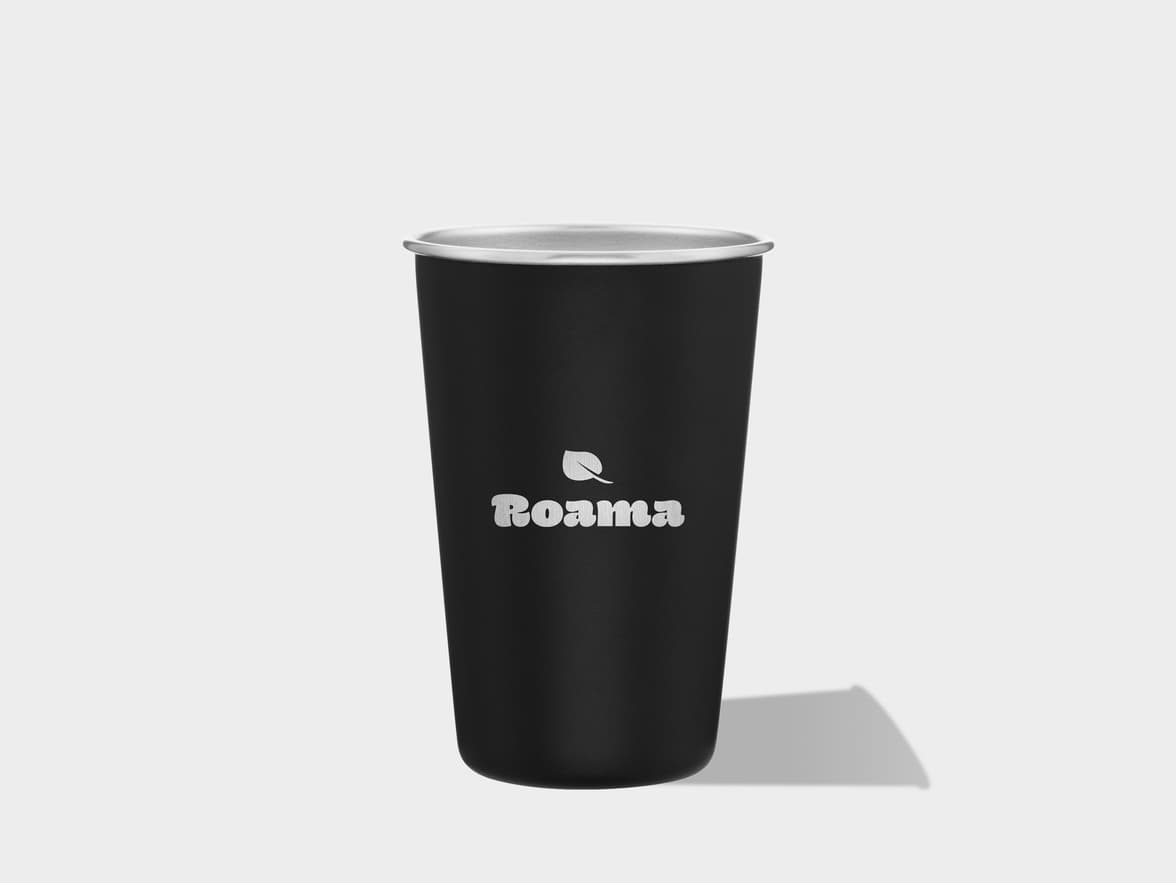 A black matte tumbler with a silver rim stands centered against a light gray background, casting a shadow to its right. A white logo featuring a leaf above the word "Roama" is printed on the front.