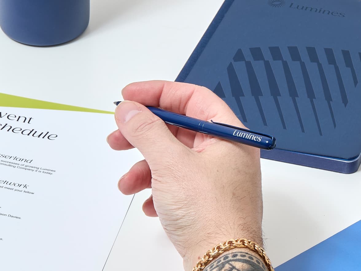 A hand holds a blue pen with "Lumines" branding, poised over a white document.