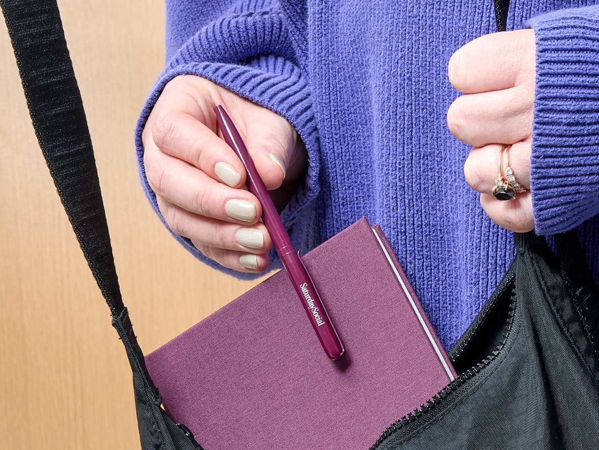 A person wearing a purple sweater holds a mulberry pen with "SaturdaySocial" branding over a mulberry notebook, inserting them into a black bag.