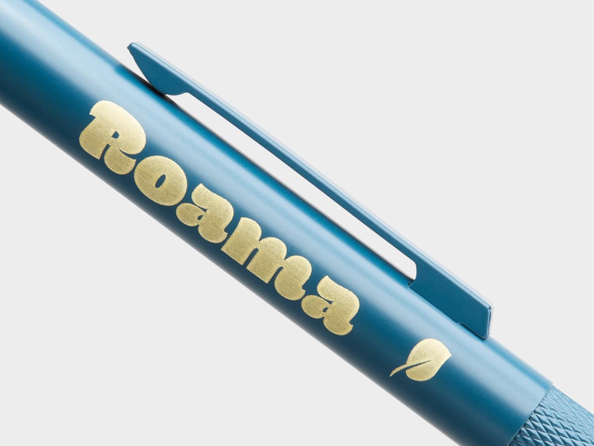 Close-up of a teal pen with the golden "Roama" logo and a golden leaf.