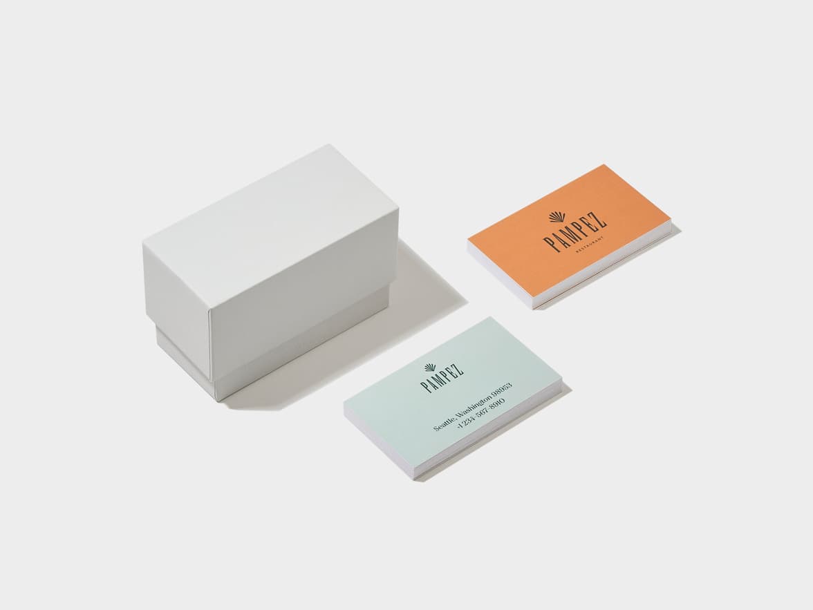 A white box next to two stacks of business cards. One stack is orange with "PAMPEZ RESTAURANT" and a palm logo. The other stack is light blue with "PAMPEZ", the palm logo, "Seattle, Washington 98953", and "(123) 567-8910".