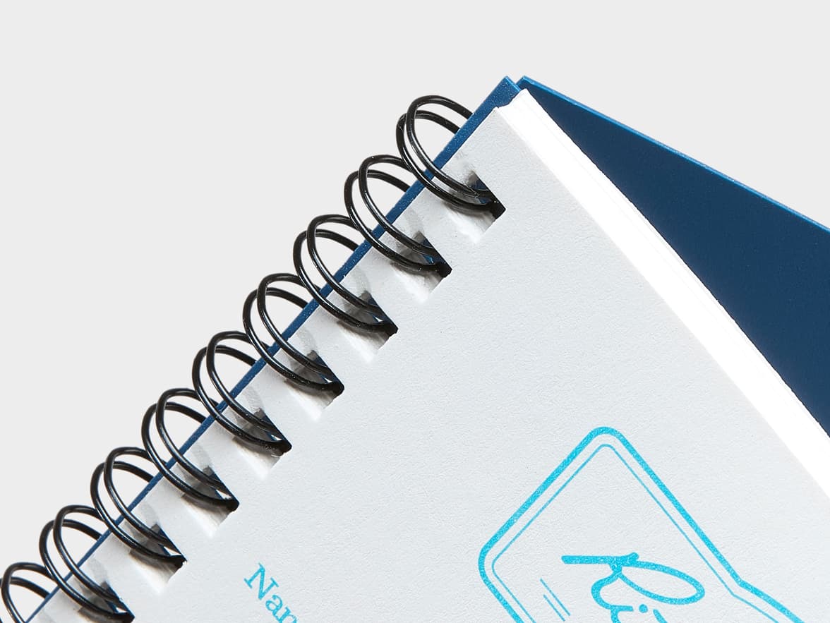 Close-up of a spiral-bound notebook with a blue cover, white textured pages, and black wire binding, showing part of a blue "Nan Ri" logo.