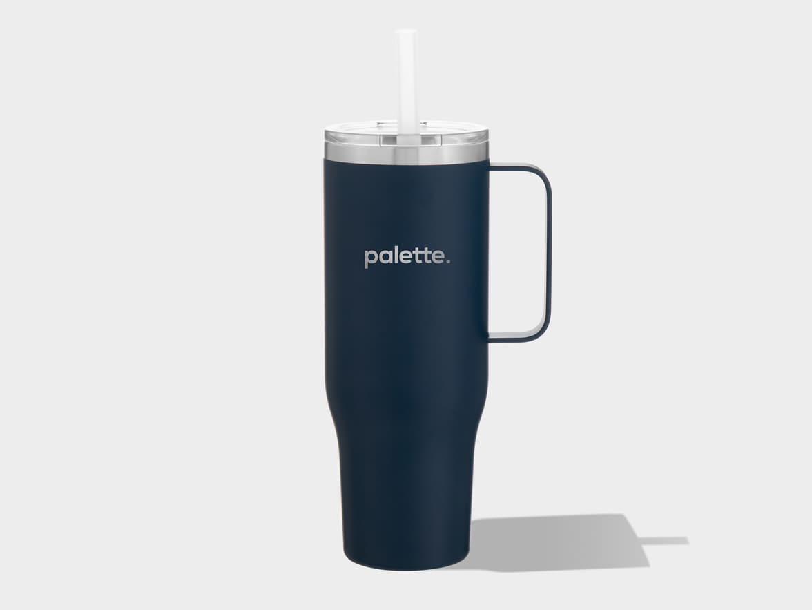 A dark blue insulated travel mug with a handle, silver rim, clear lid, and white straw, standing upright on a light gray background. The word "palette." is printed in light gray on the front of the mug.