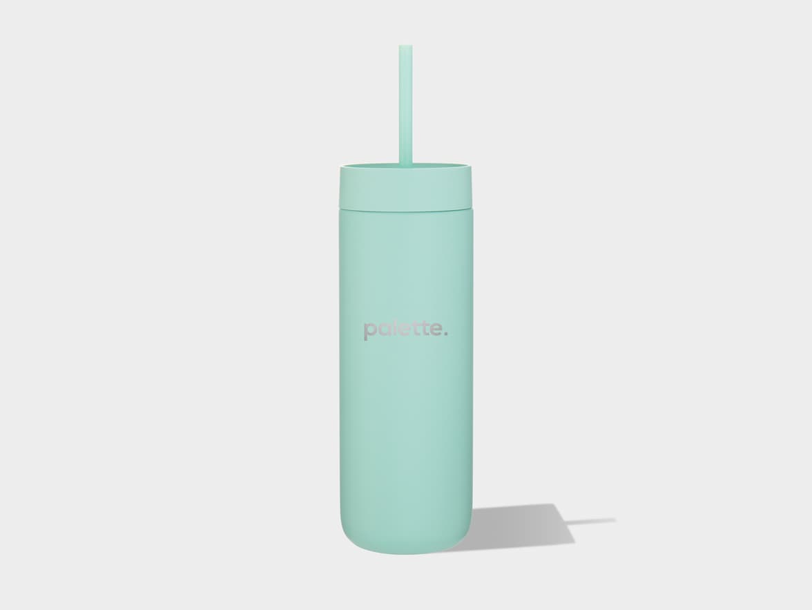 A light mint green tumbler with a straw stands upright on a light grey surface, casting a shadow to its right. The word "palette." is printed in silver letters on the side of the tumbler.