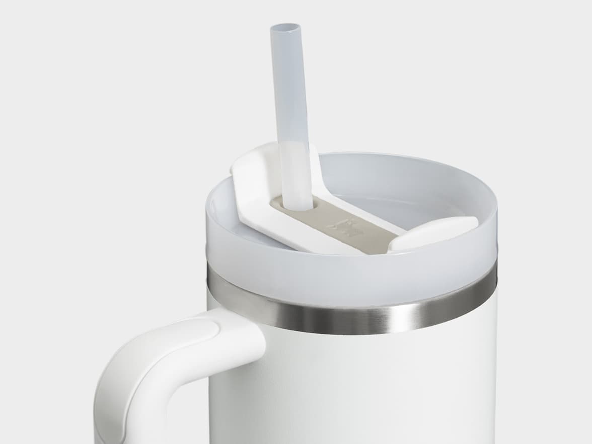 A close-up of a white insulated mug with a handle, featuring a clear straw extending from a translucent lid. The lid has a gray section with an embossed logo and is separated from the mug's white body by a silver ring.