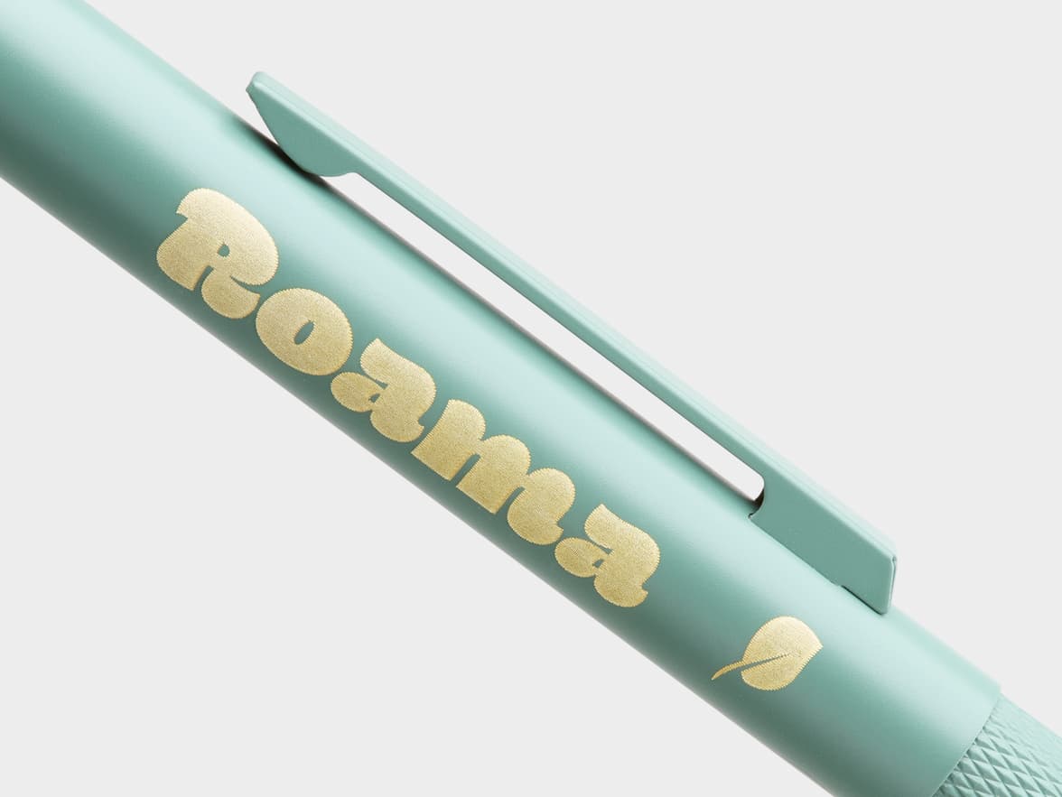 A mint green pen with a gold, textured "Roama" branding and a leaf icon.