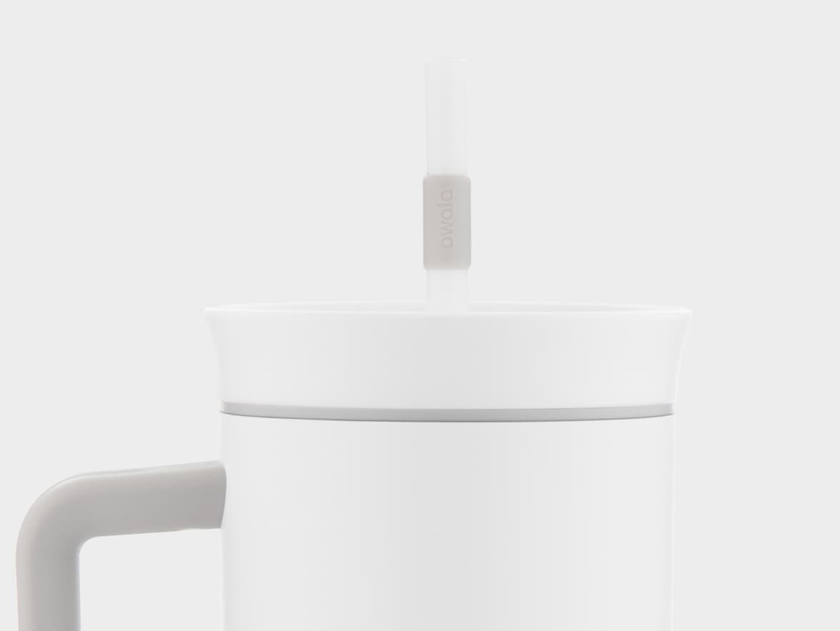 A close-up of a white Owala mug with a clear straw. The mug is white with a light grey handle visible on the left side, and it has a white lid. A translucent straw extends from the lid, featuring a light grey band with the 'owala' logo. The background is a plain light grey.