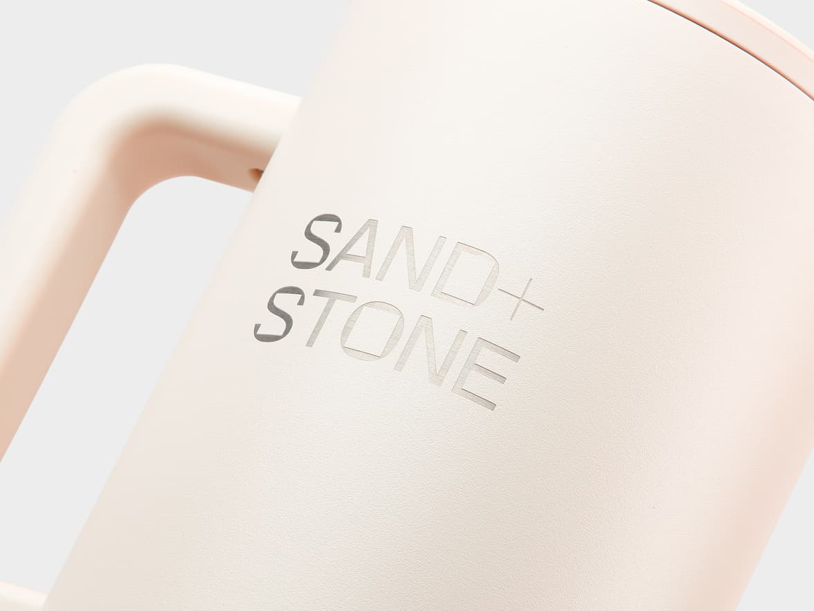 A close-up, slightly angled shot of a light, pale beige or peach-colored matte mug. The words "SAND+STONE" are debossed into the side of the mug. "SAND" is on the top line, with a small '+' symbol below and slightly to the right, followed by "STONE" on the bottom line. The debossed letters appear darker due to the recessed shadow. Part of the curved handle is visible on the left side of the mug