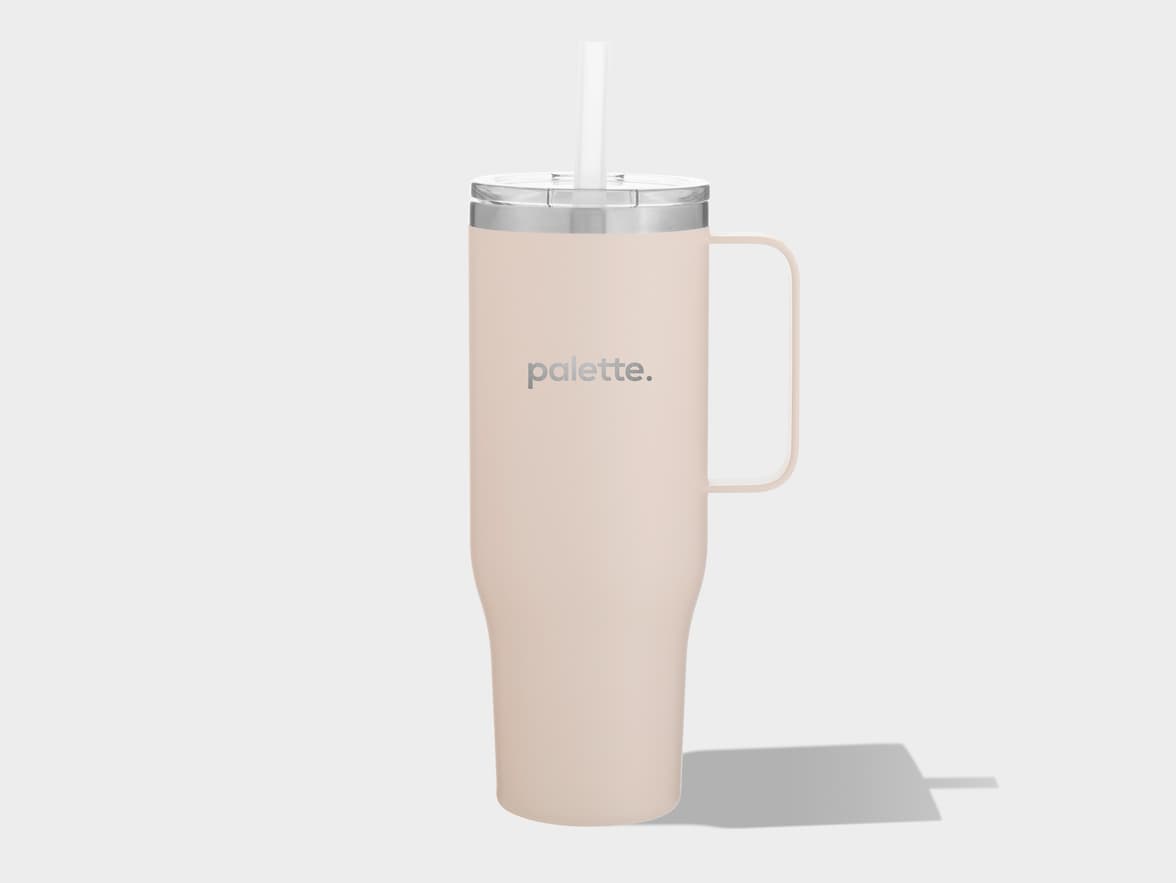 A tall, pale pink insulated tumbler with a handle on the right side and a transparent lid with a white straw extends from the top. The word "palette." is printed in gray on the front of the tumbler. It casts a long, dark gray shadow to the right on a light gray background.