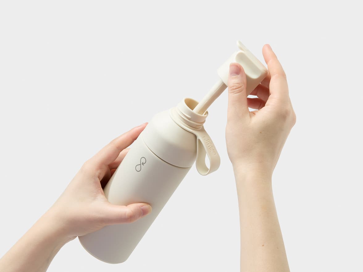 A person's hands open a cream-colored Ocean Bottle water bottle, revealing a straw-like component inside.