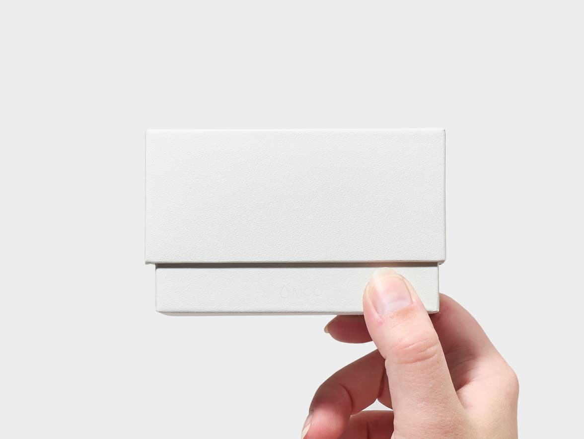 A person's hand holds a small, textured white rectangular box with the "MOO" logo subtly embossed on its lower front.