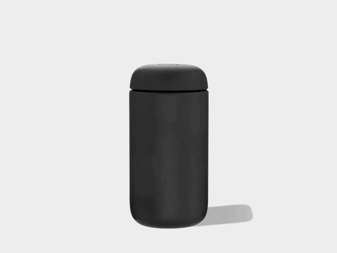A matte black thermal mug with a sleek, minimalist design stands upright on a plain light gray background. It has a cylindrical body with rounded top and bottom edges, and casts a subtle shadow to its right.