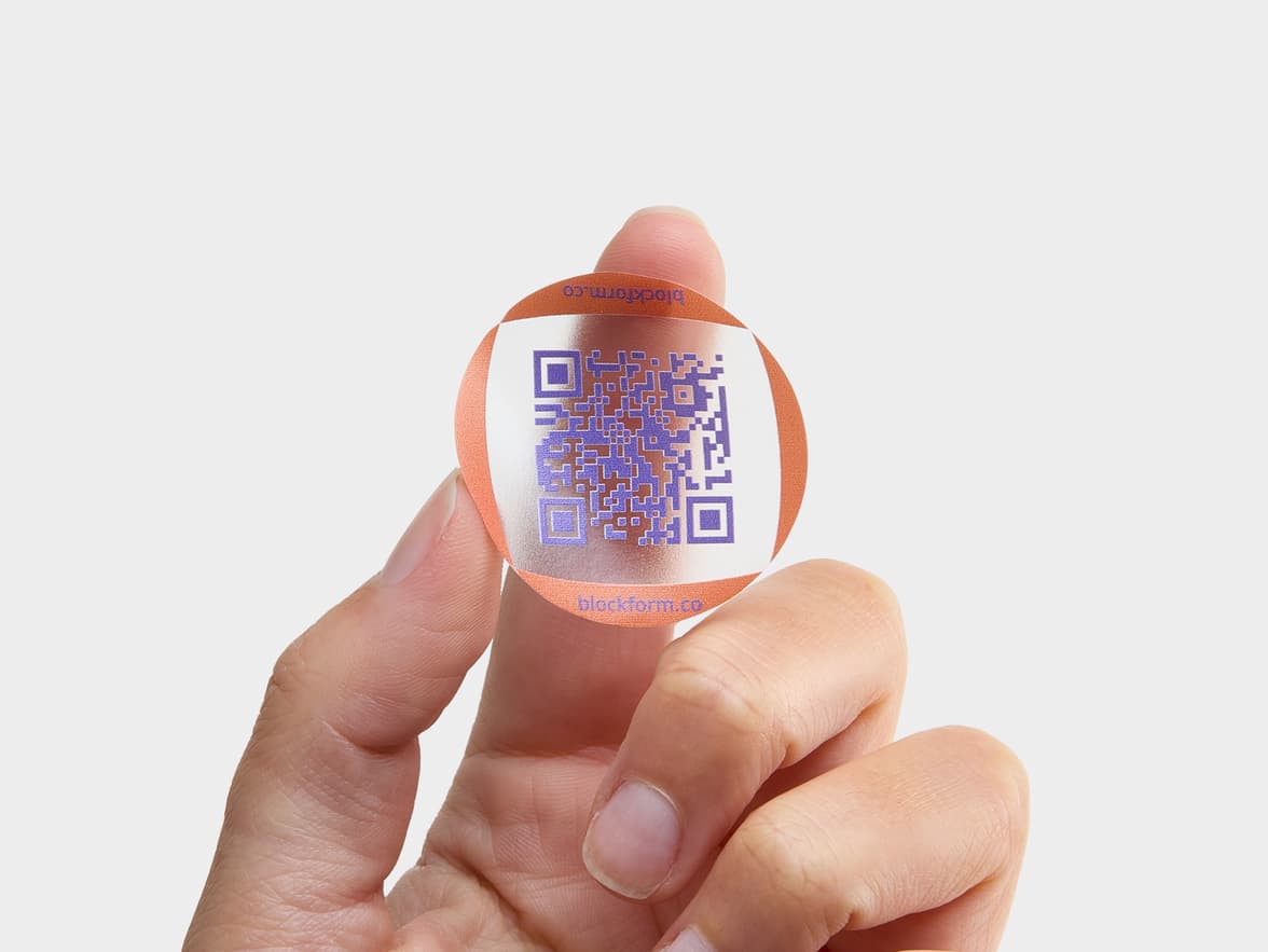A hand holding a customized clear round sticker with a purple QR code printed on it.