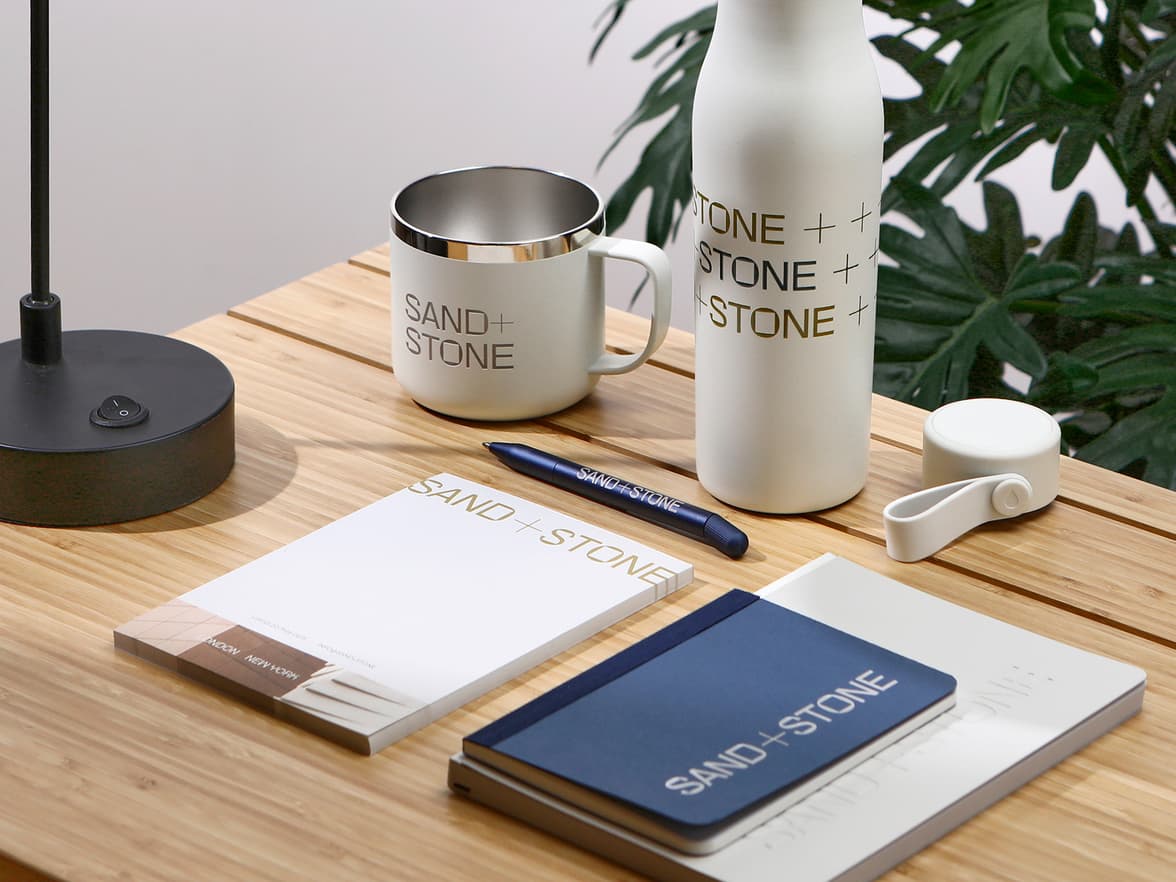 A collection of branded merchandise placed on a wooden desk including drinkware, notebooks and a pen.