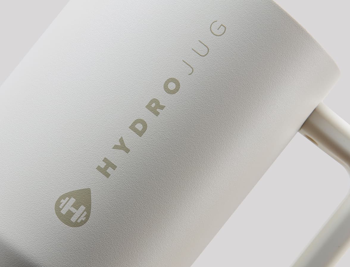 A close-up, slightly angled shot of a white matte mug. The words HYDROJUG TRAVELLER are debossed into the Tumbler